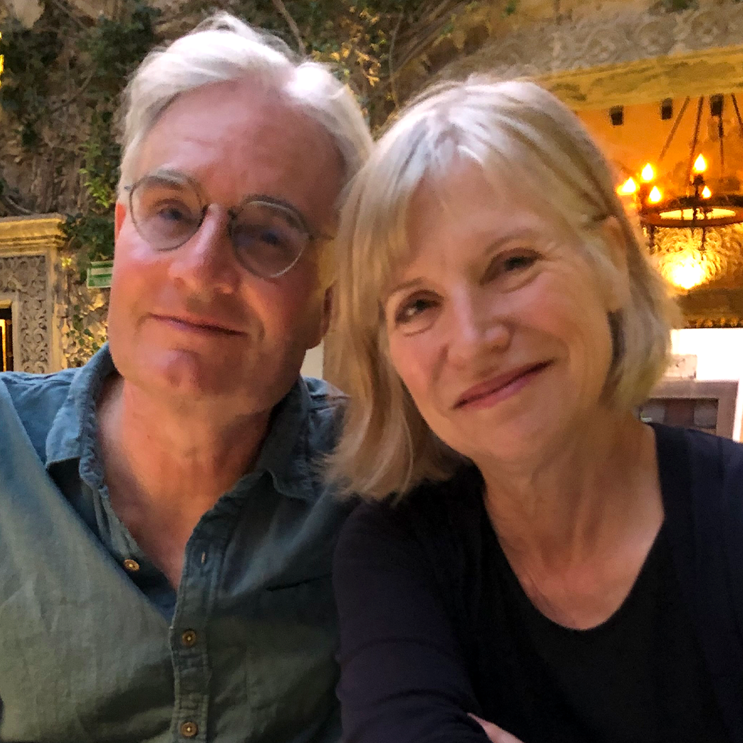 Two people sitting close together at a restaurant table with menus, a glass, and a chandelier-lit courtyard behind the        m