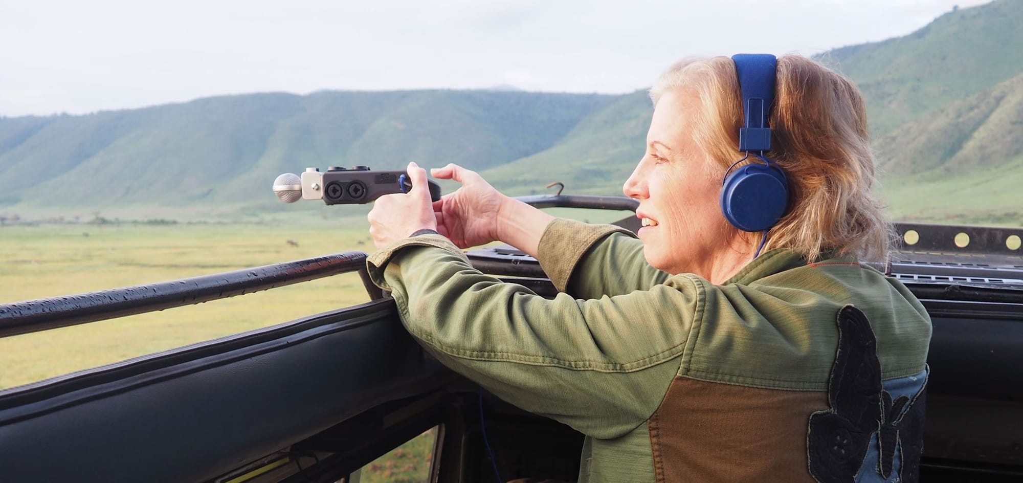 Person wearing blue headphones and an olive jacket records audio with a handheld device from an open vehicle overlooking a        green                                                                                                                                          valley and mountains  