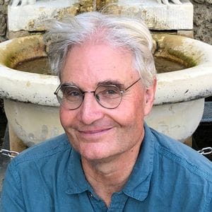 Person with gray hair and round glasses smiling in front of a stone fountain outdoors