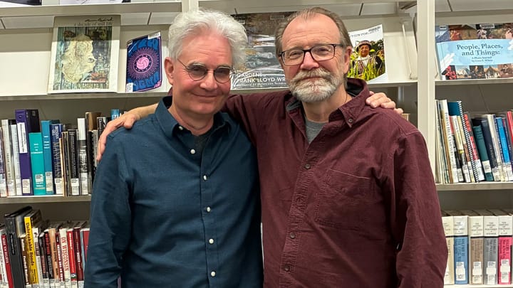 Steve Paulson and George Saunders with arms around each other's shoulders smile in front of bookshelves filled with Wisconsin-themed books. 